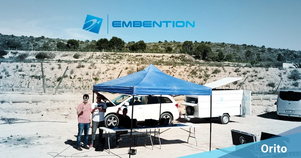 A field test setup with several people standing under a blue tent, in front of a vehicle and trailer, with the "Embention" logo prominently displayed. The scene takes place in a semi-arid, rural environment labeled "Orito."