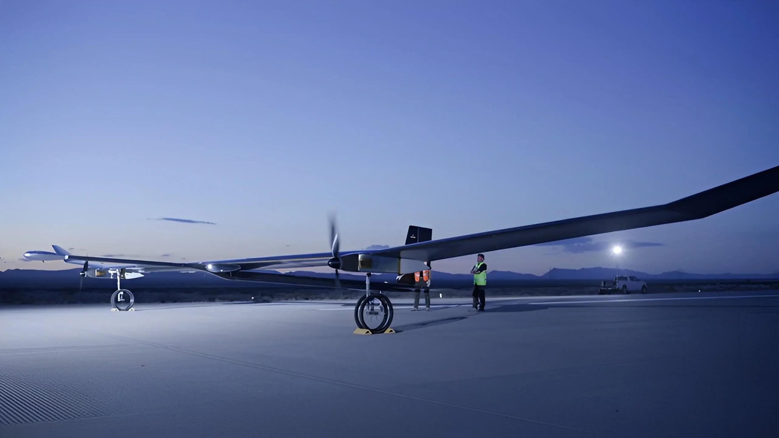 phasa-35-bae-systems-hibrido-dron Dawn scene on an airstrip with a long-winged drone preparing for flight, assisted by ground crew under a rising moon.