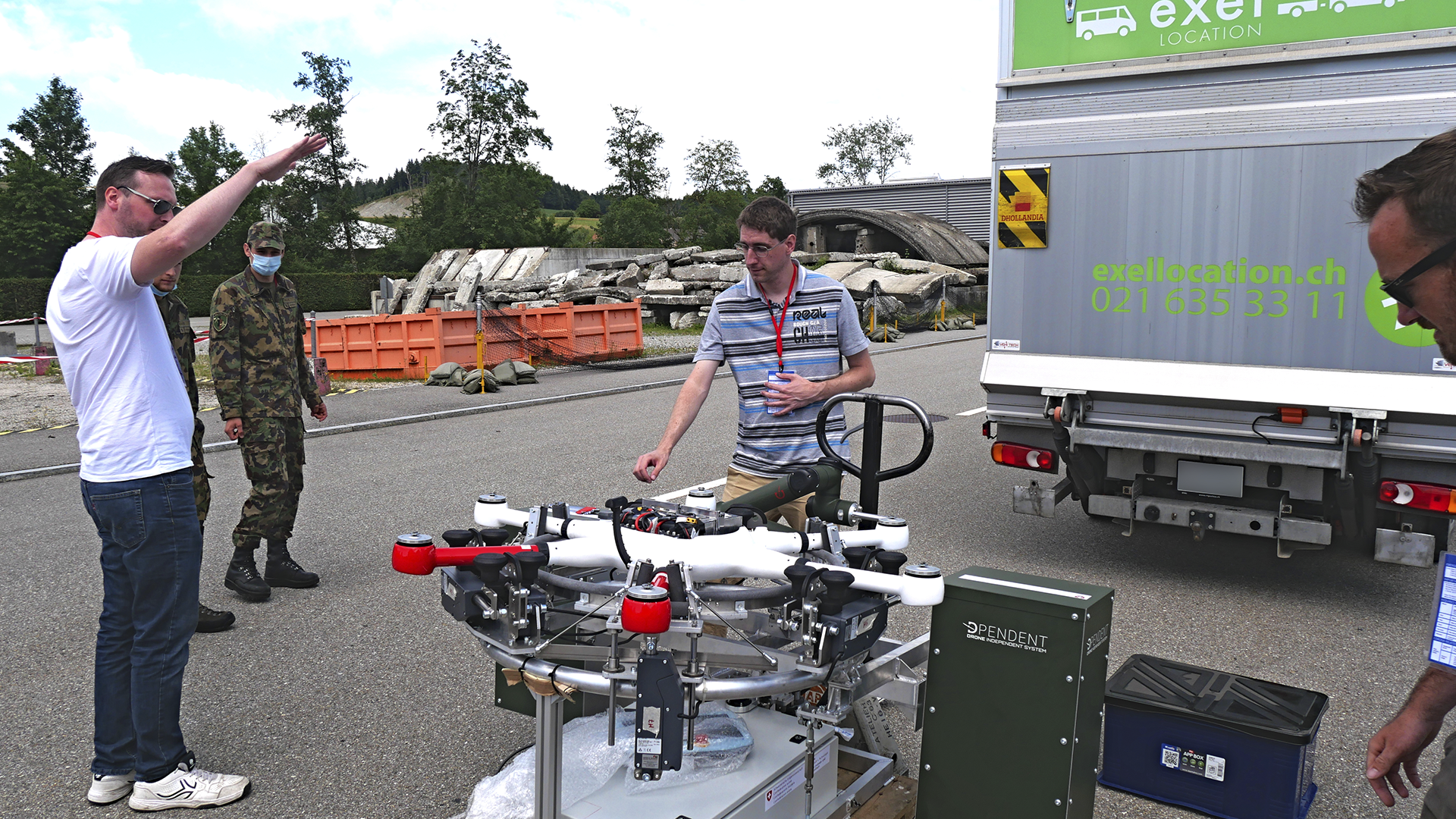 Dpendent - Unmanned Drone Ground Handling Station (UGHS) 3 A group of individuals, including a soldier and two men, interact around a drone ground handling station outdoors, while a delivery truck is parked in the background.