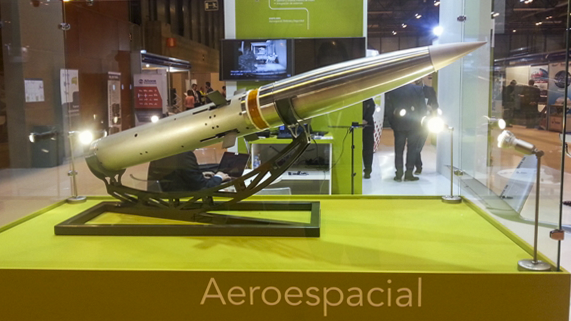 Everis - Miura Precision-guided missile on display in a glass case at an exhibition, featuring its sleek metallic design and mounted on a stand, with background activity and signage visible.