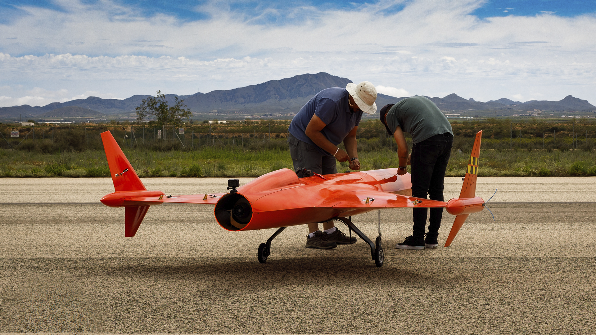 Falcon02 Two technicians inspecting and preparing a bright red unmanned aerial vehicle (UAV) on a runway with mountains in the background.